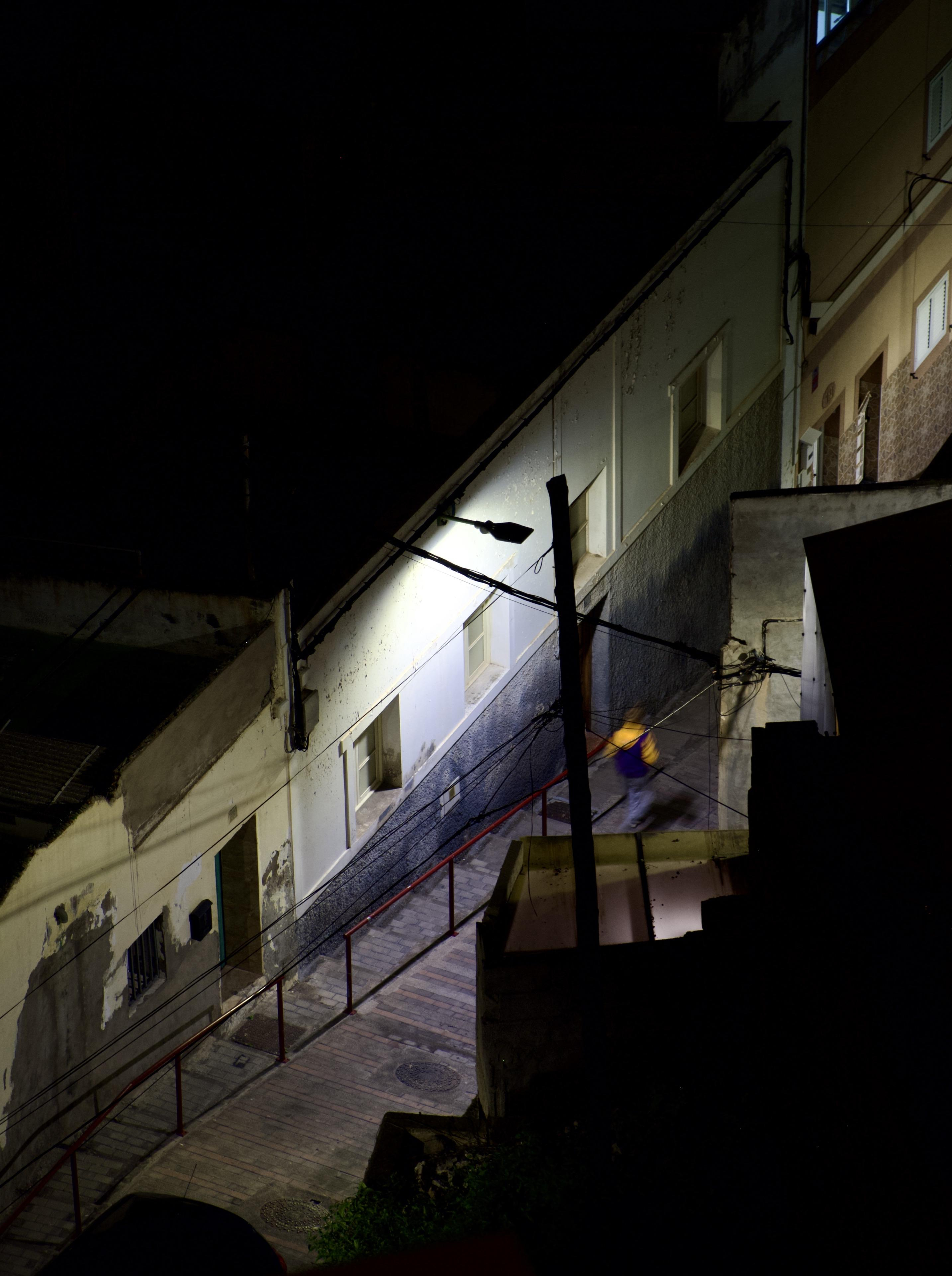 A nighttime aerial view of a narrow urban alleyway or pedestrian street, flanked by weathered multi-storey residential buildings. A single street lamp illuminates a section of the scene, casting a sharp pool of light against the white-rendered walls. Overhead wires and cables stretch across the alley, attached to a wooden utility pole. A red metal handrail runs along the sloped walkway. A motion-blurred figure wearing a yellow top is visible mid-stride on the path. The upper portion of the image is largely in darkness.