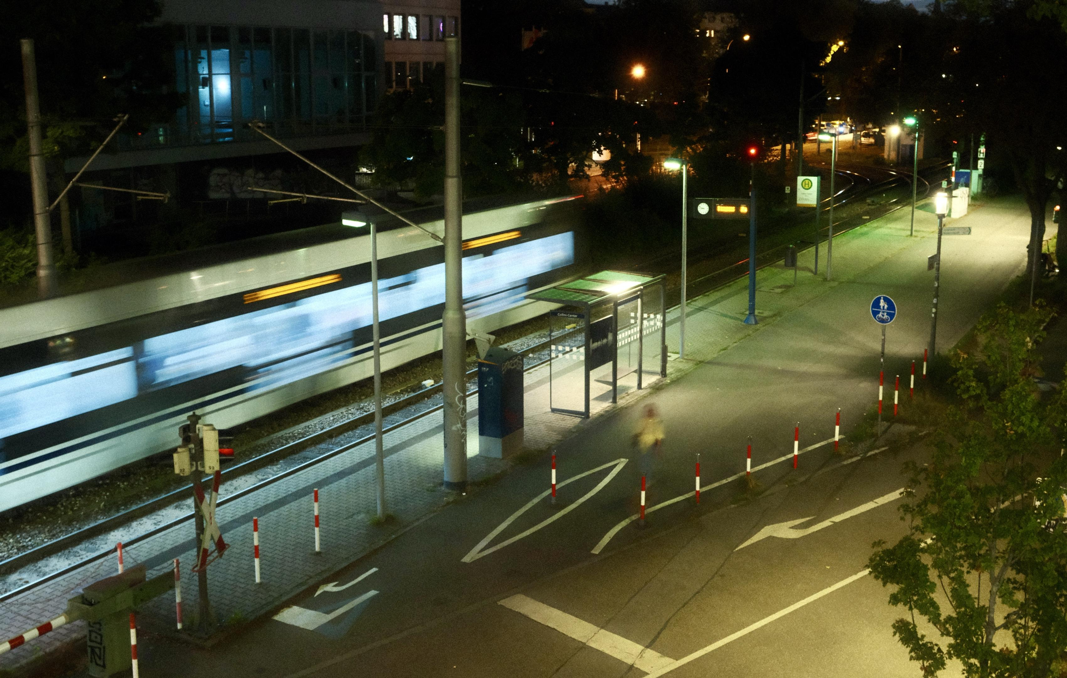 Night scene of a moving train at an urban train station, captured with motion blur. The station has a lighted platform with a bench and signage, surrounded by streetlights and trees. A pedestrian in motion is blurred on a path beside a road marked with traffic signs and flanked by red-and-white traffic poles.